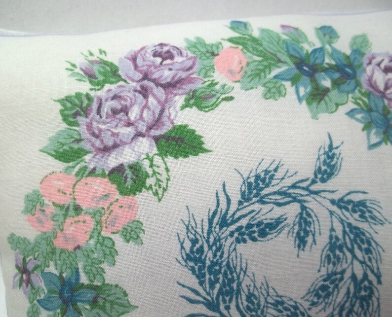 Puede incluir: Una funda de almohada blanca con un dise&ntilde;o floral que presenta rosas rosas y moradas, hojas verdes y flores azules. El dise&ntilde;o tambi&eacute;n incluye una corona de espigas de trigo en color azul verdoso.