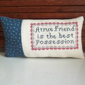 May include: A rectangular pillow with a blue floral patterned fabric on one end and a cream-colored fabric on the other. The cream section features cross-stitched text within a decorative border that reads, "A true friend is the best possession."