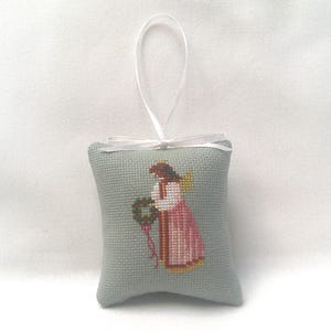 May include: A small, light blue, cross-stitched pillow ornament featuring an angel holding a wreath. The angel wears a pink dress with gold accents and has brown hair and yellow wings. A white ribbon is attached for hanging.