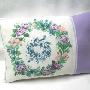 Puede incluir: Una almohada blanca con un borde lavanda presenta un dise&ntilde;o de corona floral con flores rosas y moradas, hojas verdes y una gavilla de trigo azul en el centro.