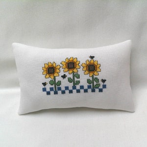 May include: A white pillow with a cross-stitch design of three yellow sunflowers with black centers and green stems. The sunflowers are arranged in a row with a blue and white checkered pattern below them.