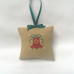 May include: A beige cross-stitched Christmas ornament in the shape of a pillow. The ornament features a red bell design with green accents and a green ribbon bow. A green ribbon is attached for hanging.