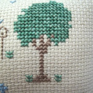 Pode incluir: Um close-up de um tecido bege com um desenho de ponto cruz verde e marrom de uma &aacute;rvore. A &aacute;rvore tem um tronco marrom e folhas verdes.