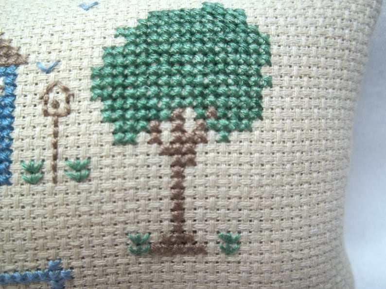 Pode incluir: Um close-up de um tecido bege com um desenho de ponto cruz verde e marrom de uma &aacute;rvore. A &aacute;rvore tem um tronco marrom e folhas verdes.