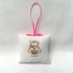 May include: A small, white, square pillow with a cross-stitched Easter bunny design. The bunny wears a yellow hat and holds a basket. A pink ribbon is attached to the top for hanging. The pillow is a decorative Easter ornament.