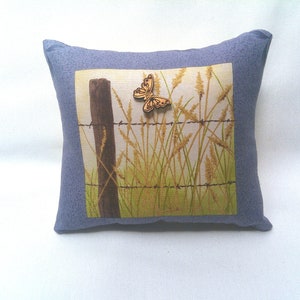 May include: A decorative pillow with a blue fabric cover and a printed image of a wooden butterfly flying over a fence with tall grass.