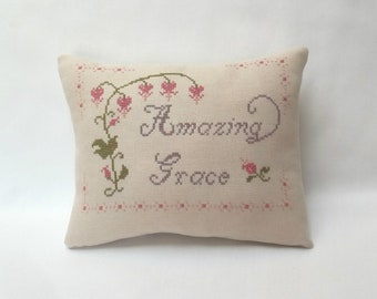 May include: A white pillow with a pink and green floral design and the words "Amazing Grace" embroidered in purple thread.