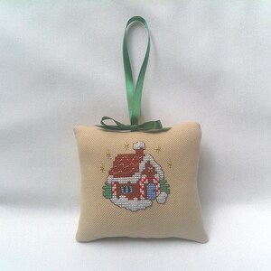 May include: A beige cross-stitch Christmas ornament shaped like a small pillow. The ornament features a red-roofed gingerbread house with white snow and green accents. A green ribbon is attached for hanging.