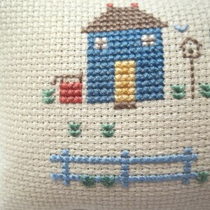 Pode incluir: Um desenho de ponto cruz de uma casa azul com uma porta amarela, um po&ccedil;o vermelho e uma cerca azul. O desenho est&aacute; sobre um fundo de tecido bege.