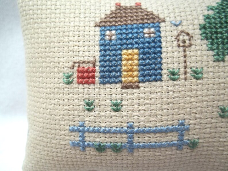 Pode incluir: Um desenho de ponto cruz de uma casa azul com uma porta amarela, um po&ccedil;o vermelho e uma cerca azul. O desenho est&aacute; sobre um fundo de tecido bege.