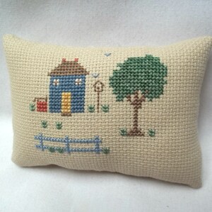 Pode incluir: Uma almofada bege com um desenho de ponto cruz de uma casa azul com uma porta amarela, uma caixa de correio vermelha, uma &aacute;rvore verde e uma cerca marrom.