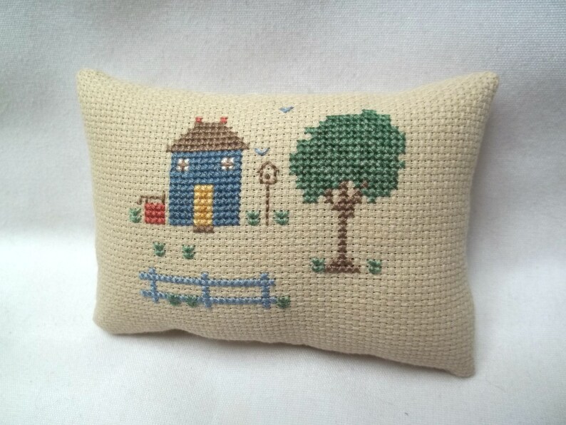 Pode incluir: Uma almofada bege com um desenho de ponto cruz de uma casa azul com uma porta amarela, uma caixa de correio vermelha, uma &aacute;rvore verde e uma cerca marrom.