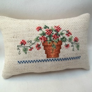 May include: A rectangular beige fabric cushion featuring a cross-stitch embroidery of a brown terracotta pot filled with red flowers and green leaves. A horizontal blue cross-stitch border runs along the bottom of the potted plant design.