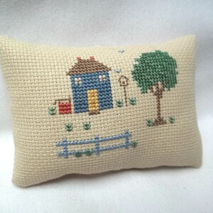 Pode incluir: Uma pequena almofada bege com um desenho de ponto cruz de uma casa azul com uma porta amarela, uma caixa de correio vermelha, uma &aacute;rvore verde e uma cerca azul.