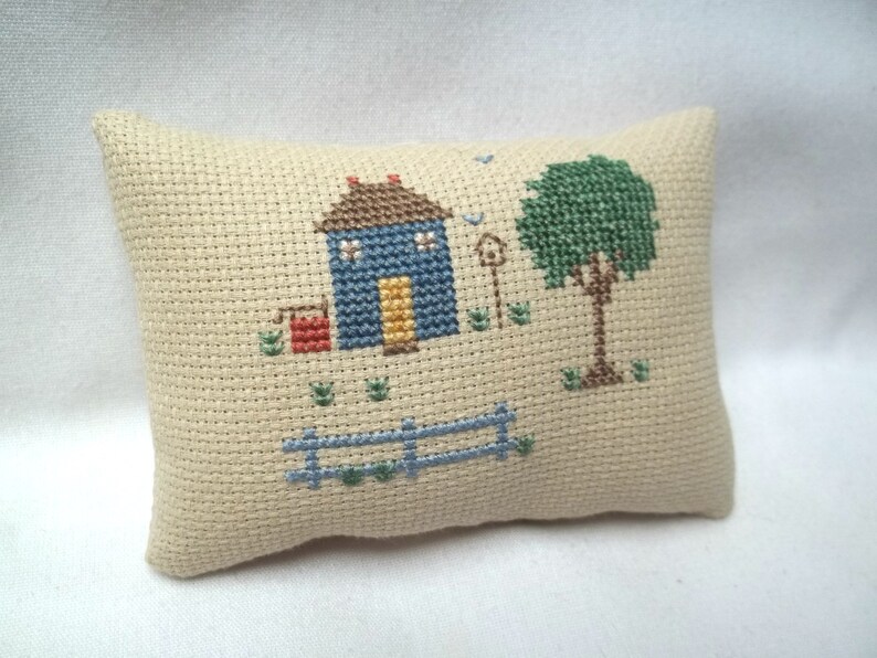 Pode incluir: Uma pequena almofada bege com um desenho de ponto cruz de uma casa azul com uma porta amarela, uma caixa de correio vermelha, uma &aacute;rvore verde e uma cerca azul.