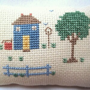 Pode incluir: Uma almofada de ponto cruz com uma casa azul com uma porta amarela, um balde vermelho, uma &aacute;rvore verde e uma cerca azul. O fundo &eacute; de tecido bege.