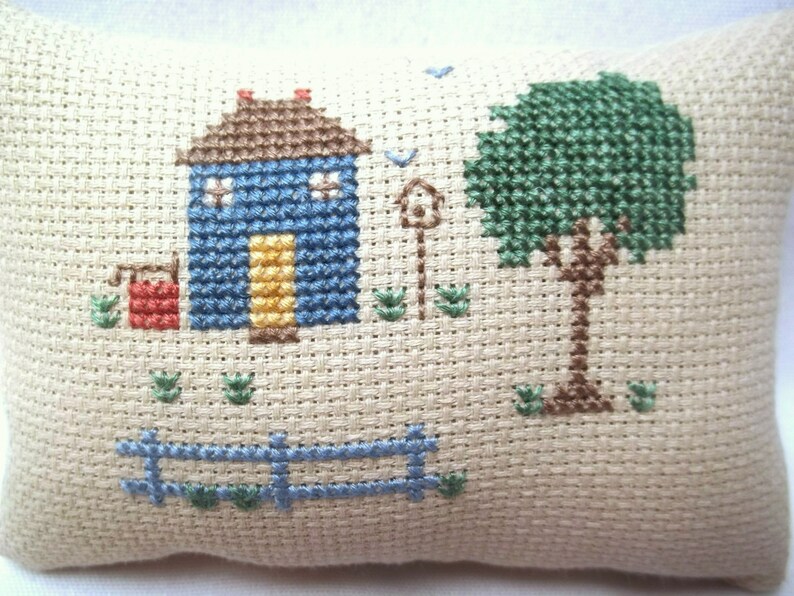 Pode incluir: Uma almofada de ponto cruz com uma casa azul com uma porta amarela, um balde vermelho, uma &aacute;rvore verde e uma cerca azul. O fundo &eacute; de tecido bege.