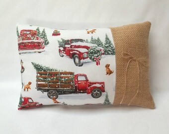 May include: Rectangular decorative pillow with a burlap side and a printed front featuring red vintage trucks, Christmas trees, and brown dogs in a snowy scene. The pillow has a neutral background and a twine bow.