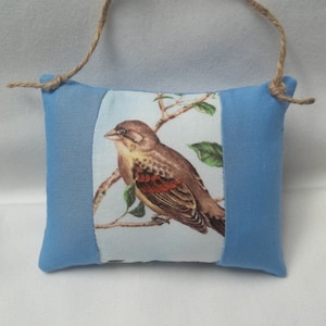 May include: A blue fabric hanging ornament with a bird and floral design. The ornament has a loop of twine at the top for hanging.