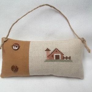 May include: A brown and beige fabric pillow with a stitched image of a red barn with a white door and a brown chimney. The pillow has two brown buttons and a brown twine loop for hanging.