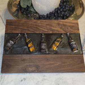 Pode incluir: Uma t&aacute;bua de servir de madeira com uma incrusta&ccedil;&atilde;o de resina cinza escuro, com cinco garrafas de whisky Jack Daniel's em miniatura. A t&aacute;bua tem um gr&atilde;o de madeira rico e escuro e est&aacute; colocada sobre um fundo desfocado com um vaso decorativo e uvas.