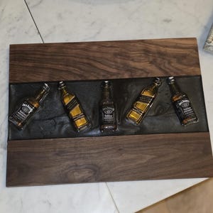 Pode incluir: Uma bandeja de madeira com cinco garrafas de whisky Jack Daniel's em miniatura embutidas em resina cinza escuro. A bandeja tem uma borda de madeira escura e formato retangular.