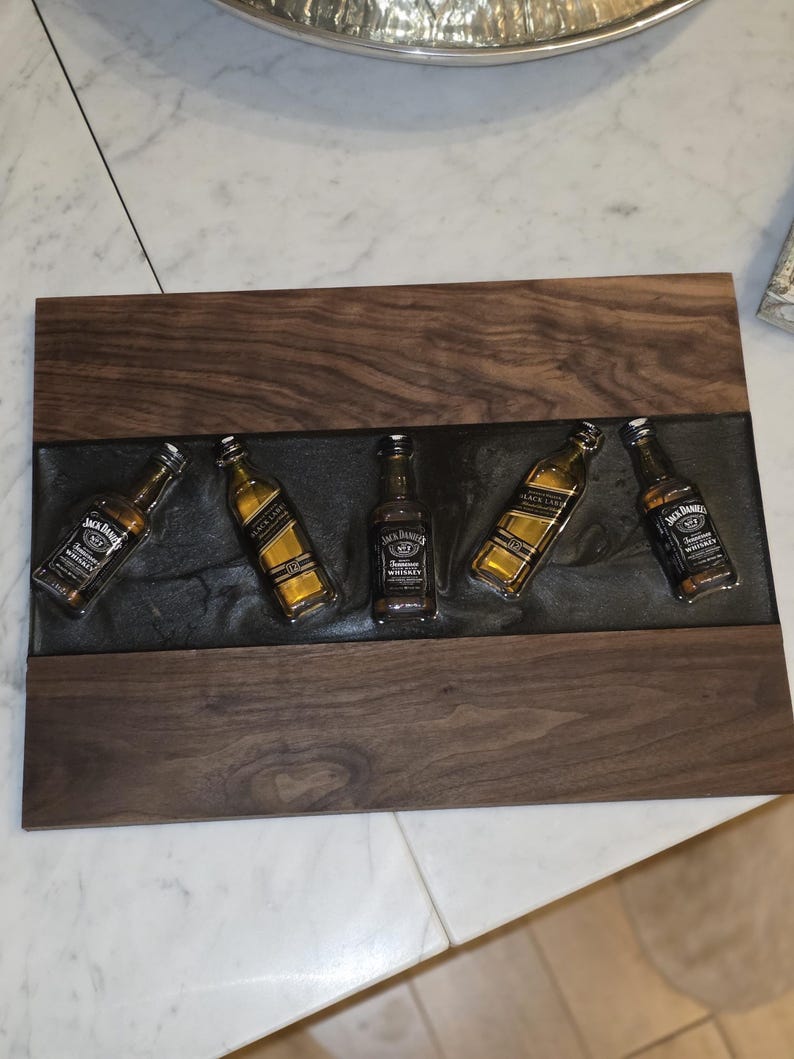 Pode incluir: Uma bandeja de madeira com cinco garrafas de whisky Jack Daniel's em miniatura embutidas em resina cinza escuro. A bandeja tem uma borda de madeira escura e formato retangular.