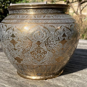 May include: A round brass vessel featuring an intricate etched pattern with silver-toned inlay. The surface displays a repeating floral and geometric motif across the body, with a wide opening at the top and a circular base at the bottom.