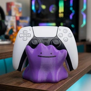 以下が含まれることがあります： 白と黒のPlayStation 5コントローラーが、紫色のDitto型のスタンドに置かれています。スタンドには笑顔があり、3Dプリント素材で作られています。コントローラーは濃い茶色の木製の表面にあります。