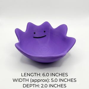 May include: A purple bowl shaped like a character with a wavy, irregular rim and a simple black line for a mouth and two black dots for eyes. The bowl has a length of 15.24 cm, a width of approximately 12.7 cm, and a depth of 5.08 cm.