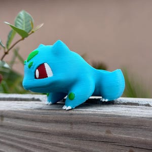 May include: A light blue Bulbasaur planter, a Pok&eacute;mon character, with red eyes, white accents, and green spots. The planter is 3D-printed and sits on a wooden surface. The planter is designed to hold small plants or succulents.