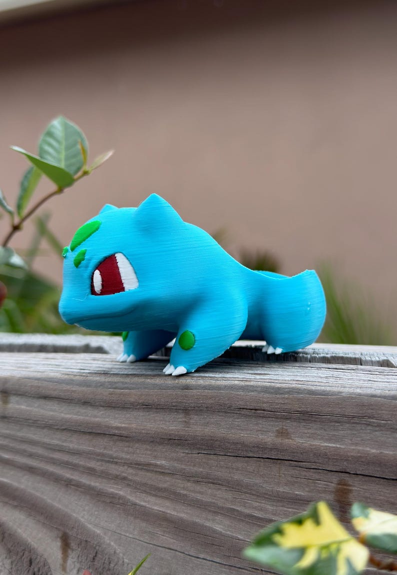May include: A light blue Bulbasaur planter, a Pok&eacute;mon character, with red eyes, white accents, and green spots. The planter is 3D-printed and sits on a wooden surface. The planter is designed to hold small plants or succulents.