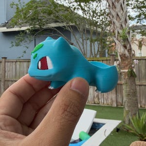 May include: A light blue 3D-printed Bulbasaur planter. The planter features Bulbasaur's iconic features, including green leaves, red eyes, and white accents. The planter is held in a hand, showcasing its size and design.