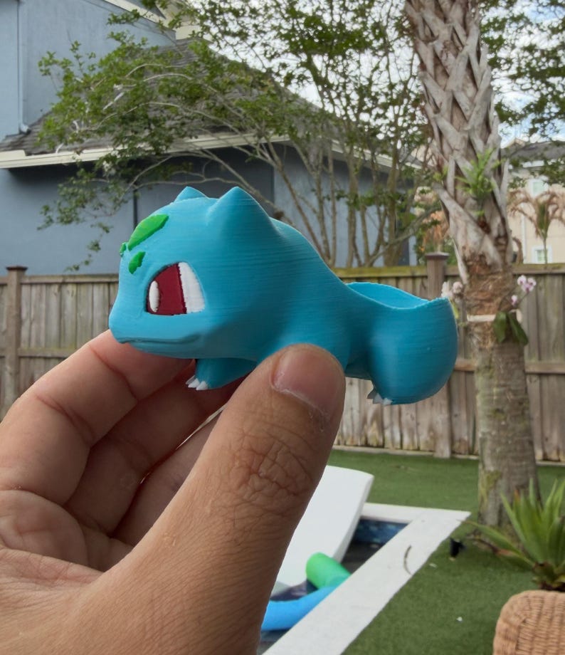 May include: A light blue 3D-printed Bulbasaur planter. The planter features Bulbasaur's iconic features, including green leaves, red eyes, and white accents. The planter is held in a hand, showcasing its size and design.
