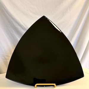 Könnte beinhalten: Eine glänzend schwarze, dreieckige Servierplatte. Der Teller hat eine glatte, reflektierende Oberfläche und wird auf einer weißen Oberfläche mit einem goldfarbenen Ständer präsentiert. Der Hintergrund ist ein weißer Stoff.