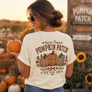 以下が含まれることがあります： 背面に「Farm Fresh PUMPKIN PATCH PICK YOUR OWN」の文字がプリントされたクリーム色のTシャツ。チェック柄、花柄、ヒョウ柄の3つのカボチャ、木製の柵、赤いリボンのデザインが描かれています。