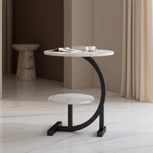 May include: A modern side table with a white marble top and a lower shelf. The table has a black, curved metal frame and a square base. A book and a cup are on the top surface.