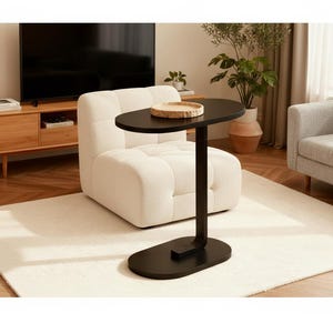 May include: A black side table with an oval top and base, connected by a curved stem. The table is positioned next to a white, tufted armchair. A wooden tray sits on the table. The room features a light-colored rug and a wooden entertainment center.