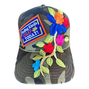 May include: Camouflage trucker hat with embroidered floral design in red, blue, orange, and pink. The hat features a patch with the text "Feeling Kinda IDGAF-ish TODAY!" in a blue and orange rectangle.