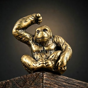 May include: A detailed, golden-colored monkey figurine in a seated pose with one arm raised. The sculpture is set against a dark background and rests on a wooden surface, showcasing intricate details.