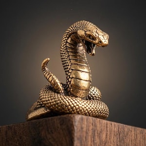 Könnte beinhalten: Eine detaillierte, goldfarbene Kobra-Statue mit offenem Maul, bereit zum Zustoßen. Die Schlange ist aufgerollt und sitzt auf einem dunklen Holzblock. Der Hintergrund ist ein Farbverlauf aus Dunkelgrau und Schwarz.