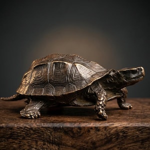 May include: A bronze-colored turtle figurine with detailed shell patterns and textured skin. The sculpture is posed on a wooden surface, showcasing its legs and head. The turtle's shell has a geometric design.