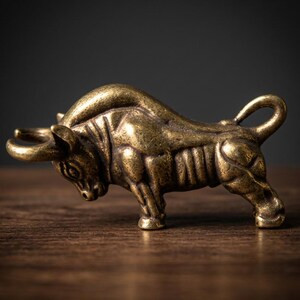 May include: A small, antique-style brass bull figurine. The bull is posed in a charging stance, with its head lowered and horns forward. The figurine has detailed musculature and a textured surface, and is set on a wooden surface.