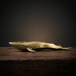 May include: A brass whale figurine, resting on a dark wooden surface. The whale is detailed with fins, a tail, and etched lines on its head. The sculpture has a polished, golden finish, and is set against a blurred, dark background.