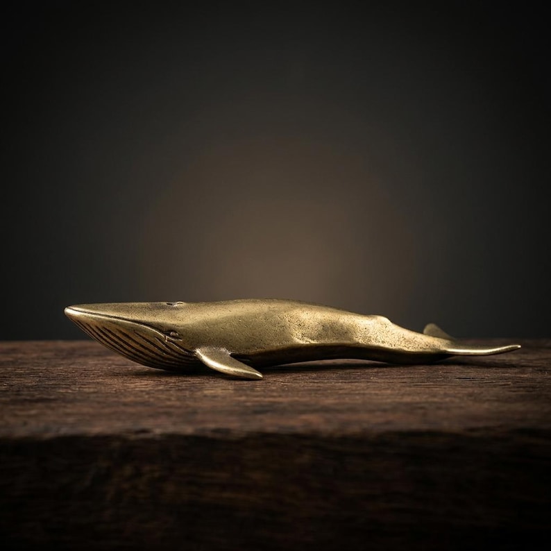 May include: A brass whale figurine, resting on a dark wooden surface. The whale is detailed with fins, a tail, and etched lines on its head. The sculpture has a polished, golden finish, and is set against a blurred, dark background.