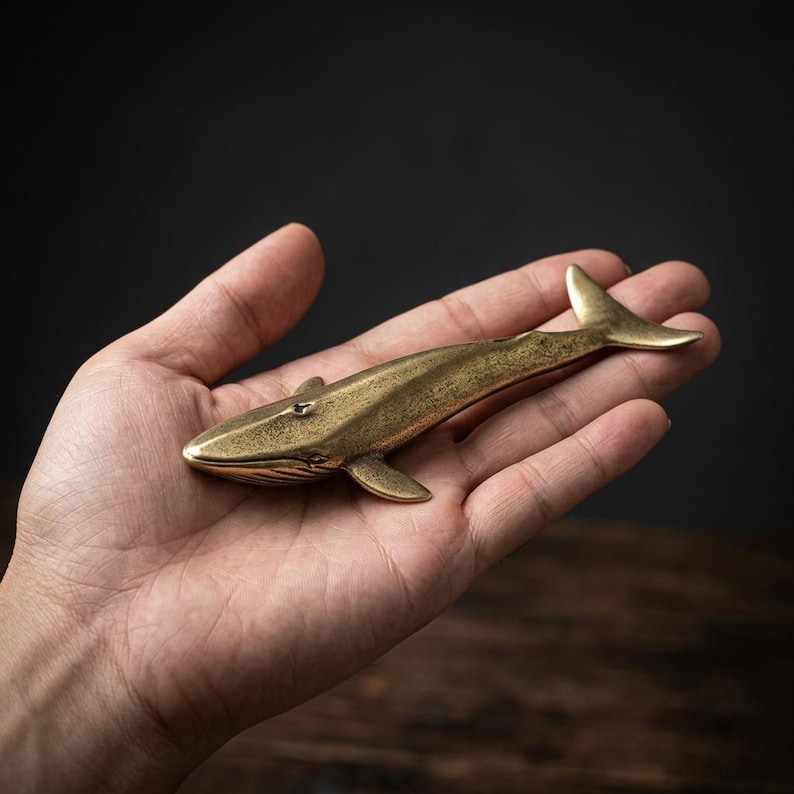 May include: A small, brass whale figurine with a detailed design. The whale is a golden color and has fins, a tail, and a small eye. The figurine is held in a person's hand, showcasing its size and craftsmanship.