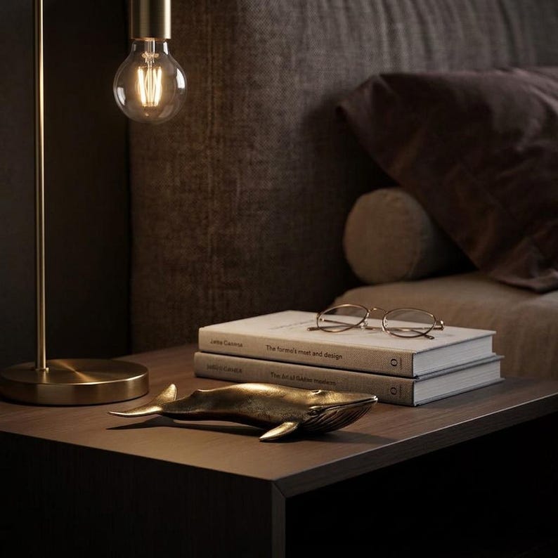 May include: A bronze whale figurine rests on a dark wooden nightstand next to two books and a pair of glasses. A gold-toned lamp with a lit bulb hangs above the nightstand, creating a warm ambiance.