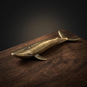 May include: A brass whale figurine with detailed fins and tail rests on a dark wooden surface. The whale is a warm gold color, with visible eye and mouth details. The background is a dark, blurred gradient.