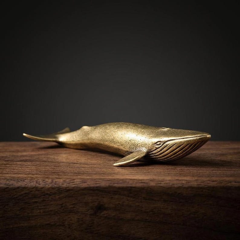 May include: A brass whale figurine with a detailed design. The whale is lying on a wooden surface. The figurine has a golden color and is a decorative item.