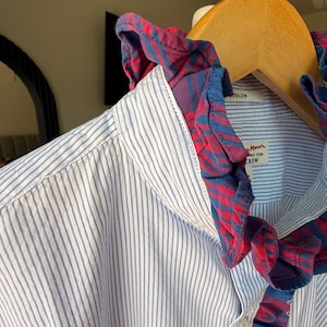 May include: A long-sleeve button-down shirt featuring a light blue and white vertical pinstripe pattern. The shirt includes a contrasting red and blue plaid ruffled trim along the collar and the front button placket. The label reads Ludlow.